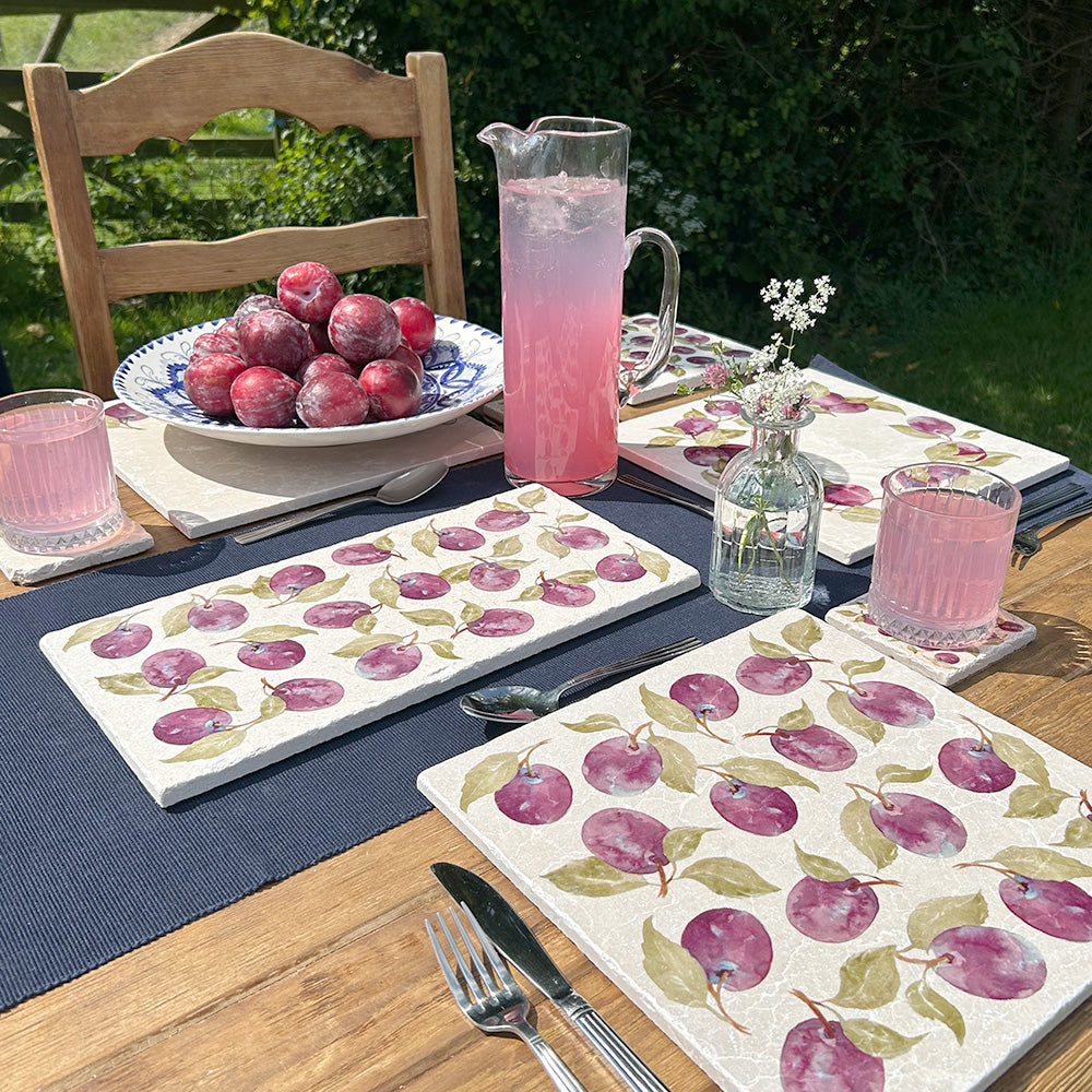 An outdoor table set with elegant bowls of fruit, vases of flowers, and drinks, placed on multipurpose marble platters featuring a watercolour plum pattern.