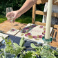 A personalised marble placemat with a plum branches wreath design, with a custom personalised message in the centre. The placemat is on a garden table set for a dinner party.