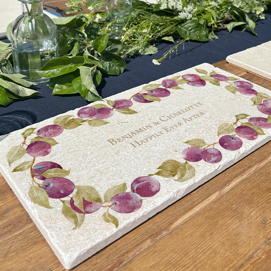 A personalised marble sharing board featuring a wreath of plums and leaves, with a bespoke message in the centre of the design. The custom sharing platter is set on a garden dining table.