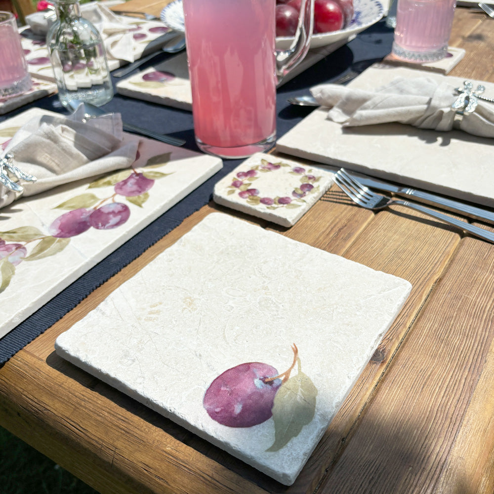 A garden table in the sunshine set with marble placemats, coasters and platters, all featuring a watercolour design of a single plum.