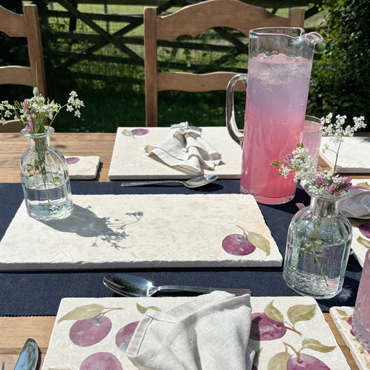 A garden table set with marble placemats and serving platters featuring a minimalistic plum watercolour design.