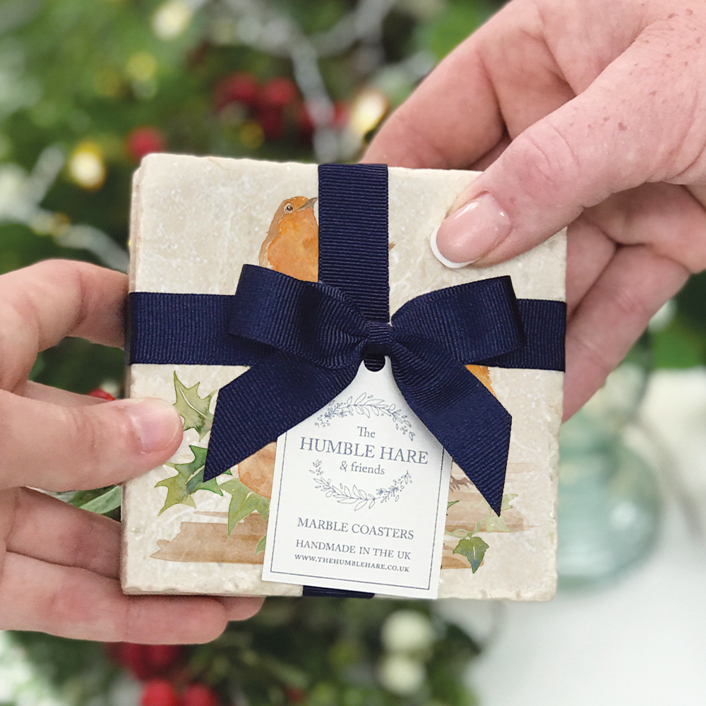 A set of 2 marble coasters featuring a Christmas robin and holly design being given as a gift. The coasters are packaged with a luxurious blue bow and gift tag.