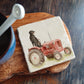 A square marble coaster with a black flat-coated retriever dog sat on a vintage red tractor. The coaster is on a kitchen work surface.