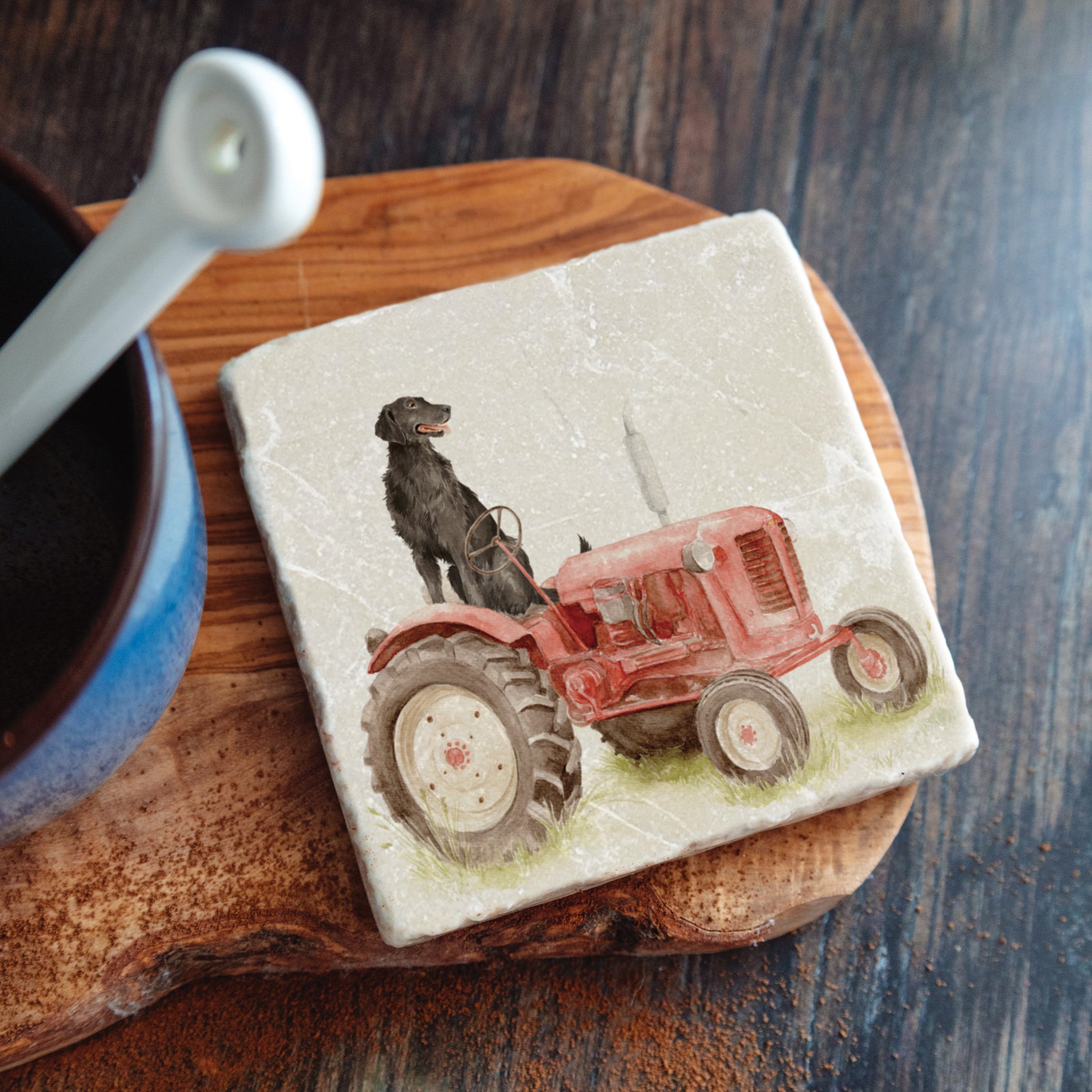 A square marble coaster with a black flat-coated retriever dog sat on a vintage red tractor. The coaster is on a kitchen work surface.