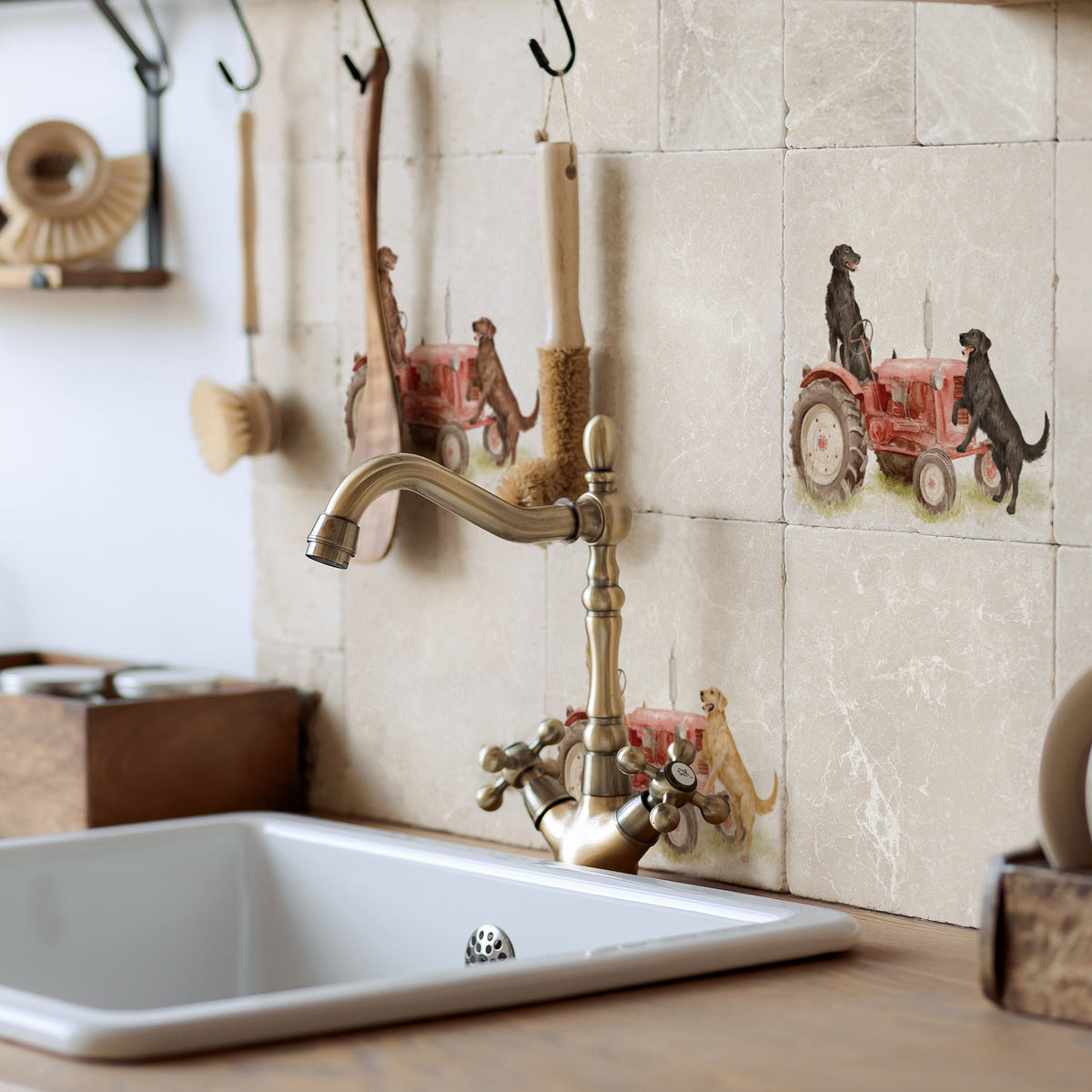 A kitchen backsplash with beige stone tiles featuring decorative illustrations of red tractors and dogs. A white rectangular sink, a brass gooseneck faucet, and rustic kitchen tools hang on hooks against the tiled wall.
