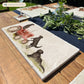 A rectangular marble platter with black retriever dogs sat on a vintage red tractor. The marble platter is sat on a farmhouse table in the garden.