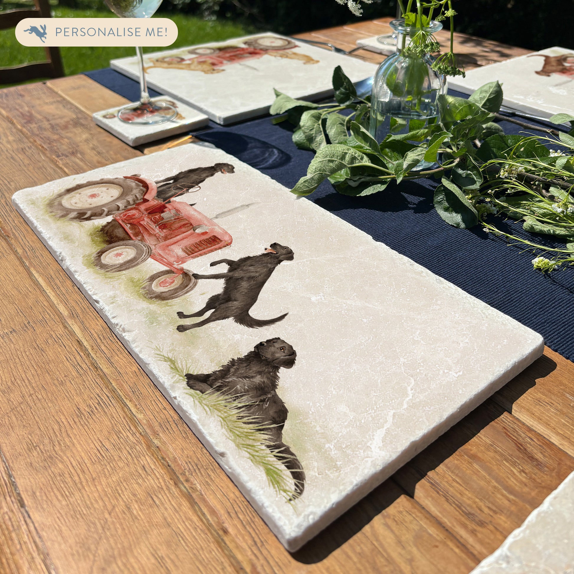 A rectangular marble platter with black retriever dogs sat on a vintage red tractor. The marble platter is sat on a farmhouse table in the garden.
