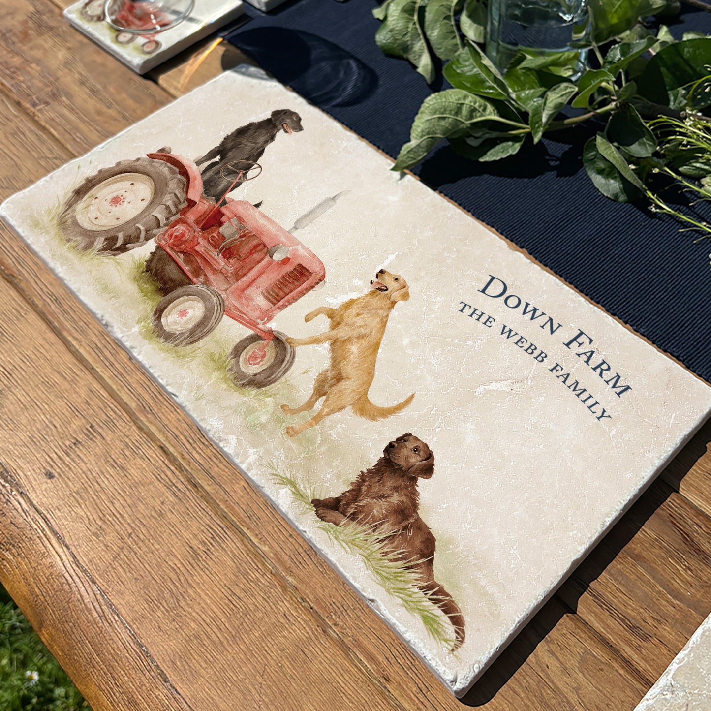 A rectangular marble platter with black, golden and chocolate retriever dogs sat on a vintage red tractor. The marble platter is personalised with custom text, and sat on a farmhouse table in the garden.