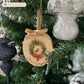 An oval marble Christmas ornament hanging on a Christmas tree featuring a robin sitting on a green wreath, with personalised text written below. The ornament has a gold velvet ribbon and bow hanging loop.