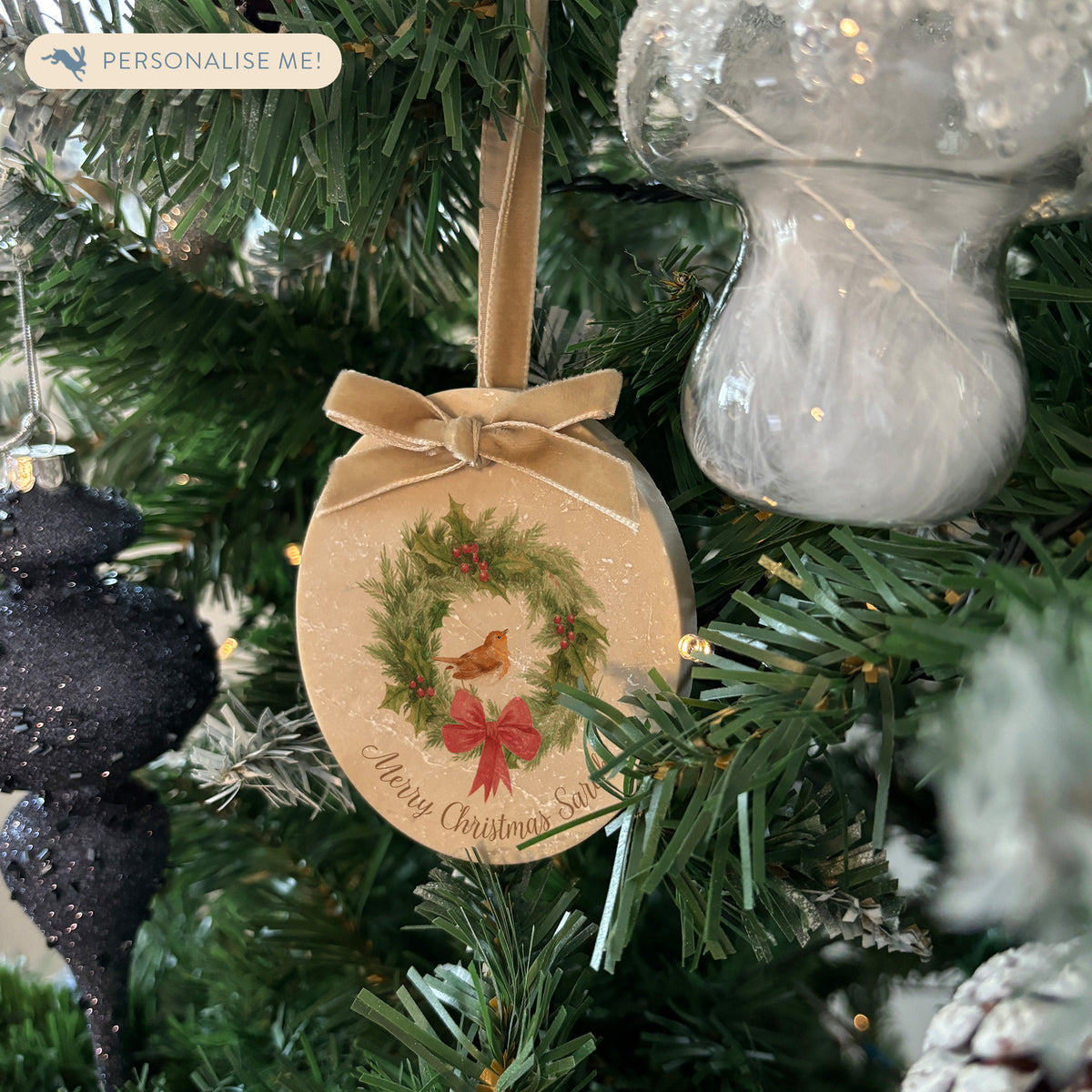 An oval marble Christmas ornament hanging on a Christmas tree featuring a robin sitting on a green wreath, with personalised text written below. The ornament has a gold velvet ribbon and bow hanging loop.