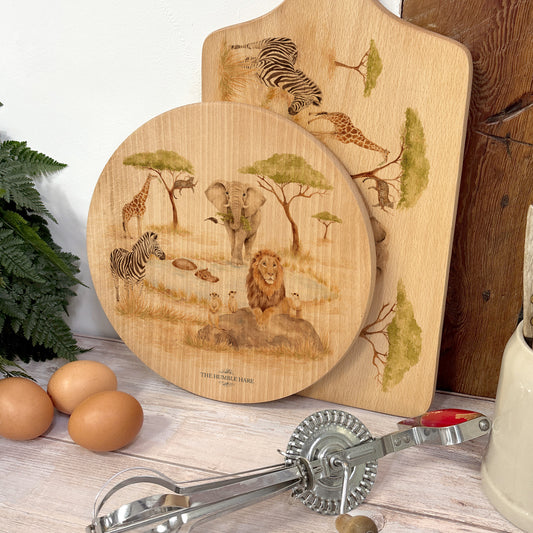 Round wooden chopping board on a kitchen worktop. The beech wood board features a watercolour design of safari animals in the African savannah.