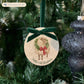An oval marble Christmas ornament hanging on a Christmas tree, featuring a sheep wearing a green wreath, with personalised text reading 'Baby's 1st Christmas' written below. The ornament has a dark green velvet ribbon and bow hanging loop.