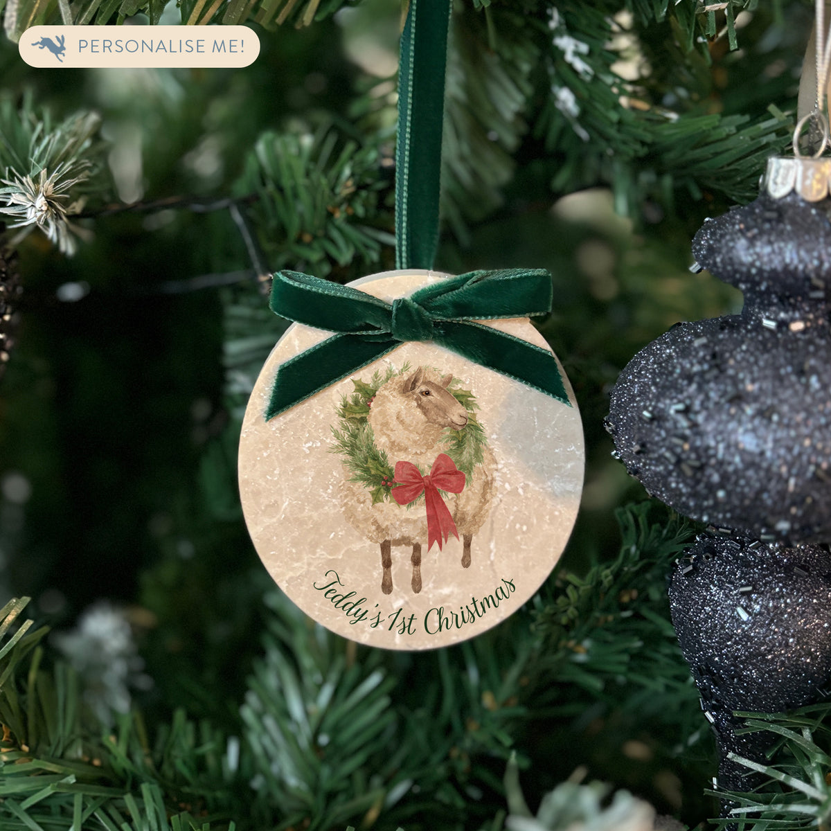 An oval marble Christmas ornament hanging on a Christmas tree, featuring a sheep wearing a green wreath, with personalised text reading 'Baby's 1st Christmas' written below. The ornament has a dark green velvet ribbon and bow hanging loop.