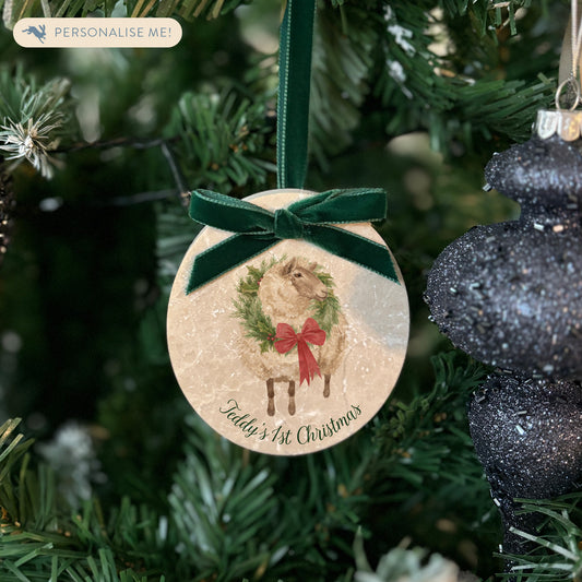 An oval marble Christmas ornament hanging on a Christmas tree, featuring a sheep wearing a green wreath, with personalised text reading 'Baby's 1st Christmas' written below. The ornament has a dark green velvet ribbon and bow hanging loop.
