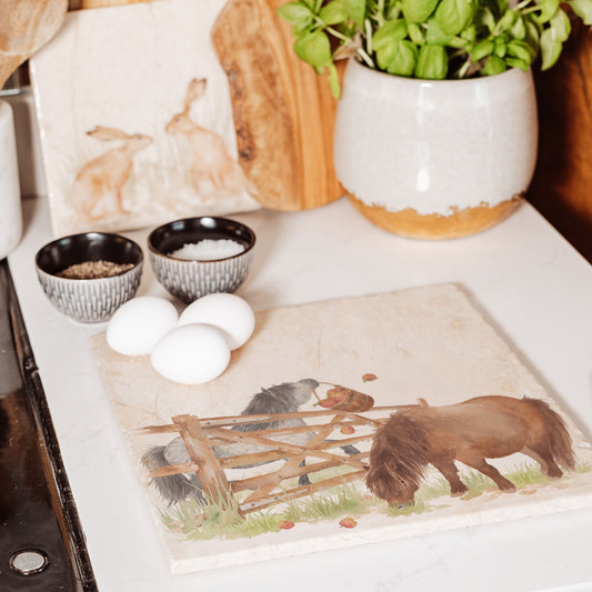 A large square multipurpose marble worktop saver, featuring a watercolour design of two cheeky Shetland ponies stealing apples.