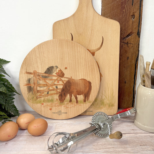 Round wooden chopping board on a kitchen worktop. The beech wood board features a watercolour design of cheeky Shetland ponies.