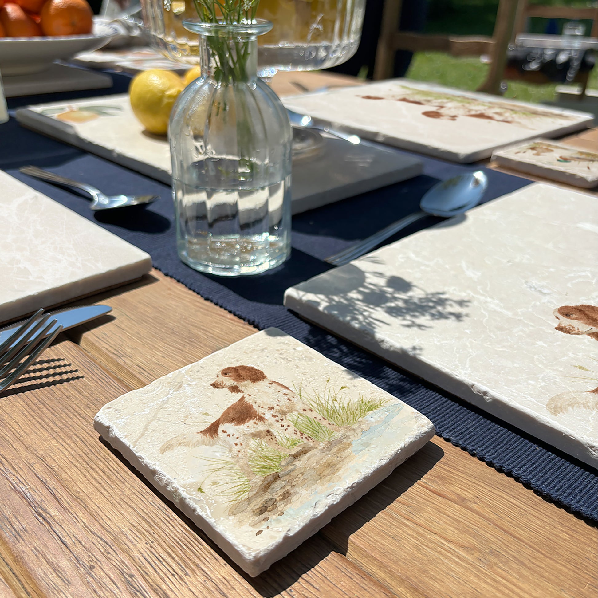 A square marble coaster with a working cocker spaniel dog by a riverbank. The coaster is set on a wooden country kitchen table.