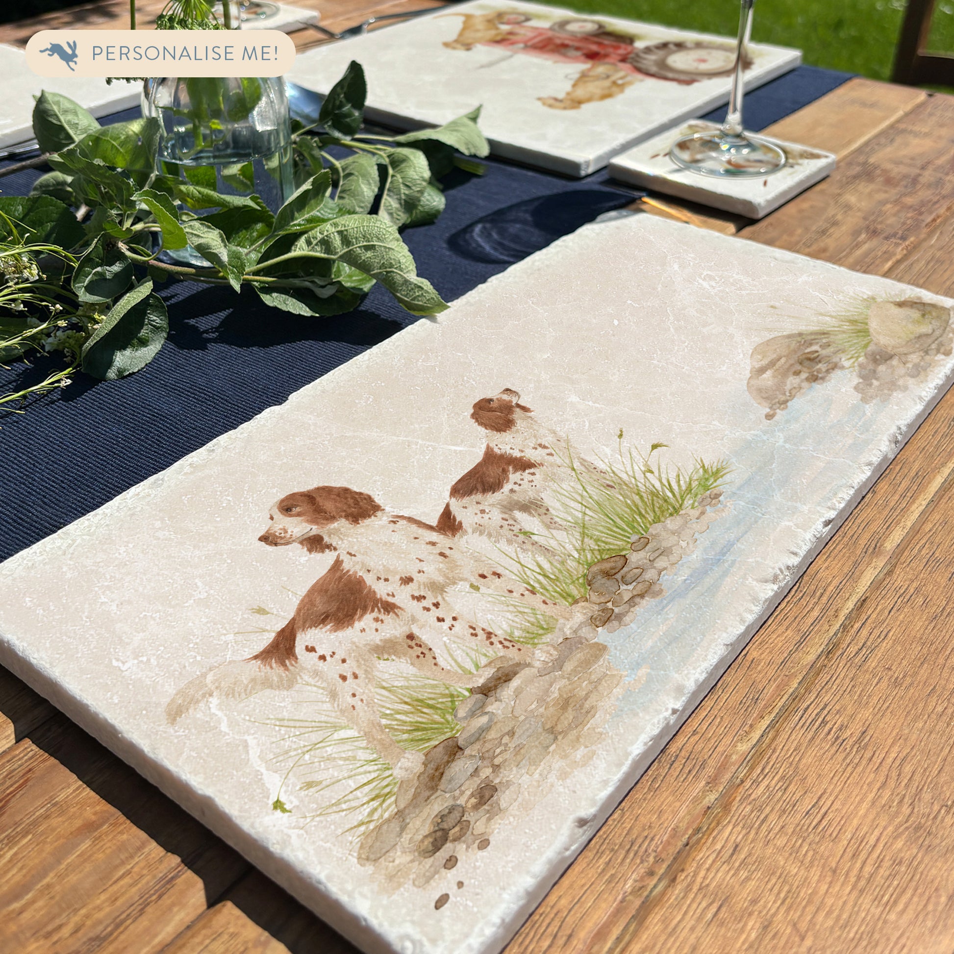 A rectangular marble sharing platter featuring a watercolour design of two working spaniel dogs by a riverbank. The platter is set on a wooden garden table.