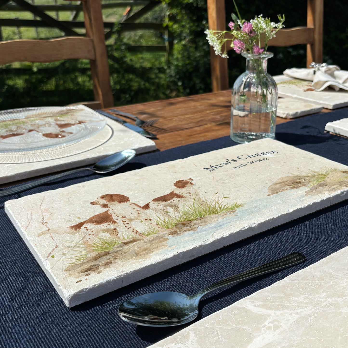 A rectangular marble sharing platter featuring a watercolour design of two working spaniel dogs by a riverbank. The platter is set on a wooden garden table.