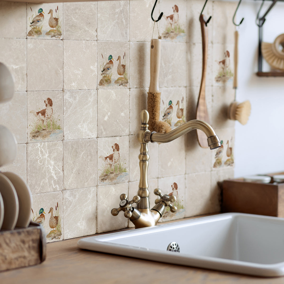 A rustic kitchen backsplash featuring square beige stone tiles with printed spaniel dog and duck illustrations, installed behind a vintage brass faucet and a white sink.