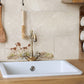 A white rectangular kitchen sink with a vintage-style brass faucet, set into a light wood countertop against a beige tiled backsplash featuring a spaniel dog illustration.