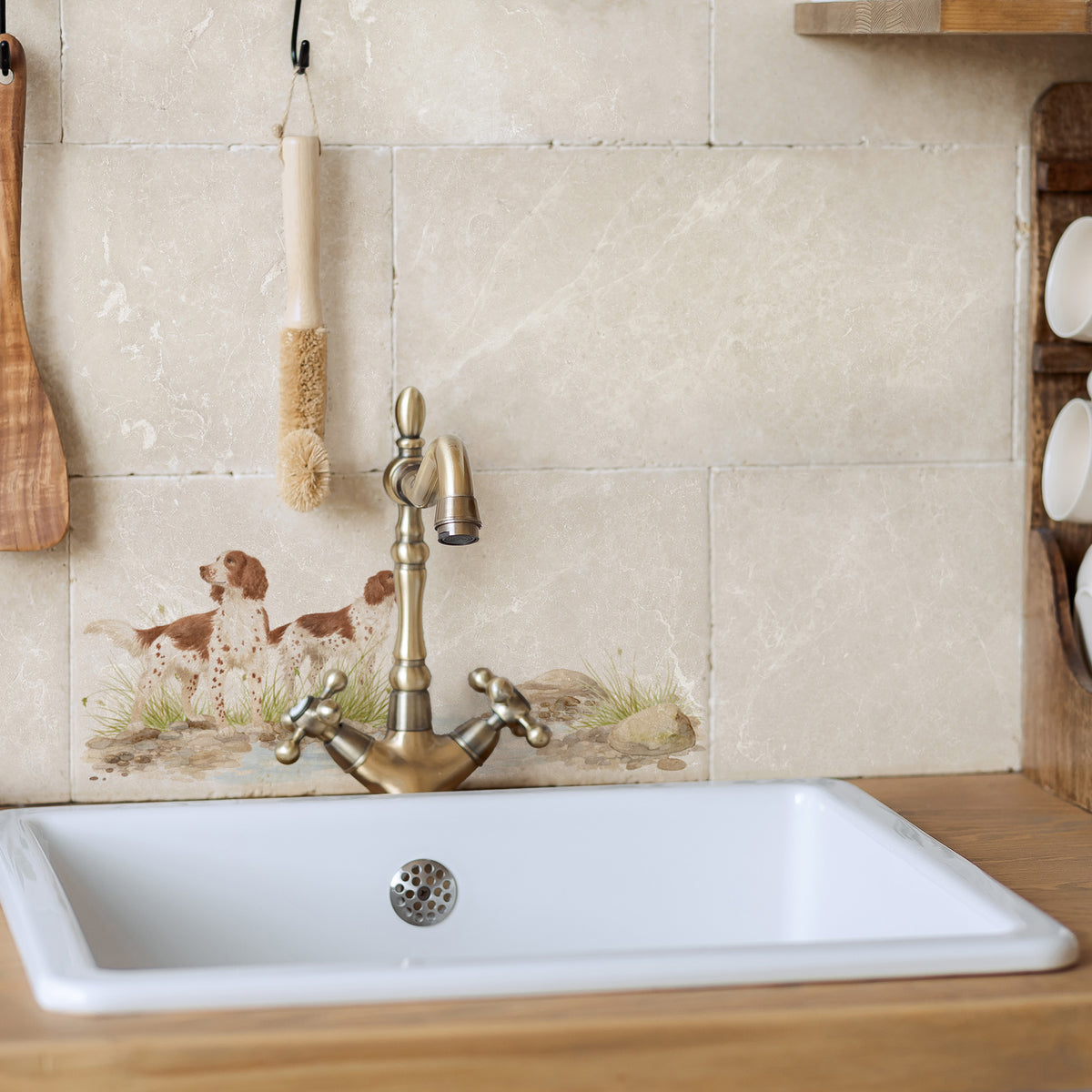A white rectangular kitchen sink with a vintage-style brass faucet, set into a light wood countertop against a beige tiled backsplash featuring a spaniel dog illustration.