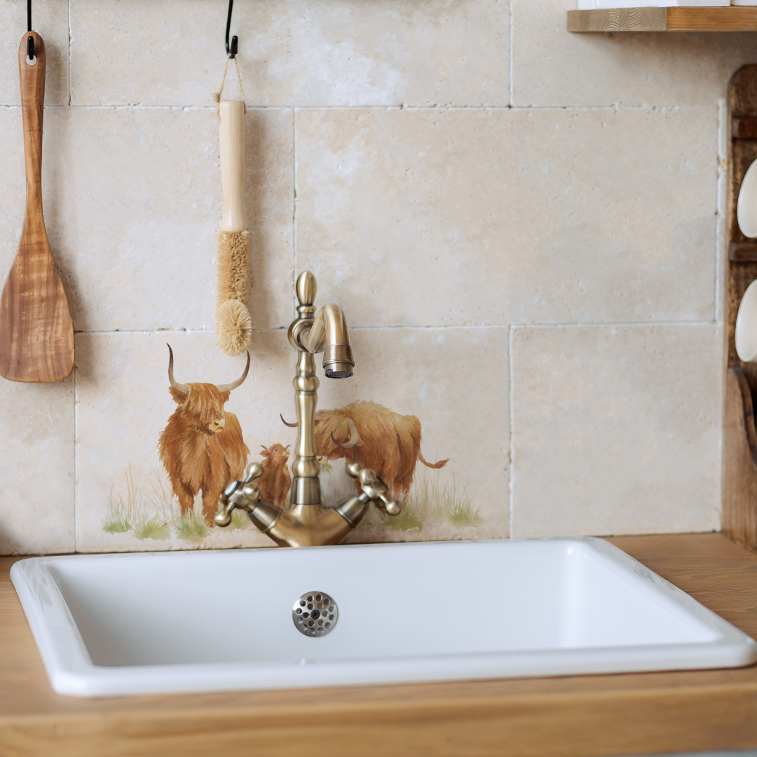 A tiled splashback behind a farmhouse kitchen sink made up of plain cream marble tiles, with a feature tile with a watercolour highland cow design.