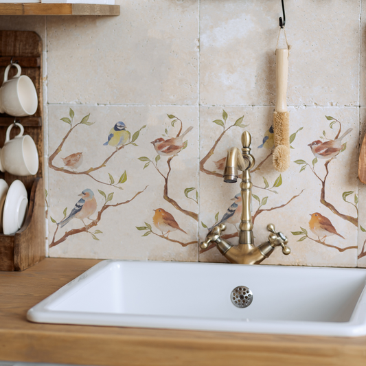 A tiled splashback behind a farmhouse kitchen sink made up of plain cream marble tiles, with a row of tiles featuring a watercolour design of garden birds on branches.