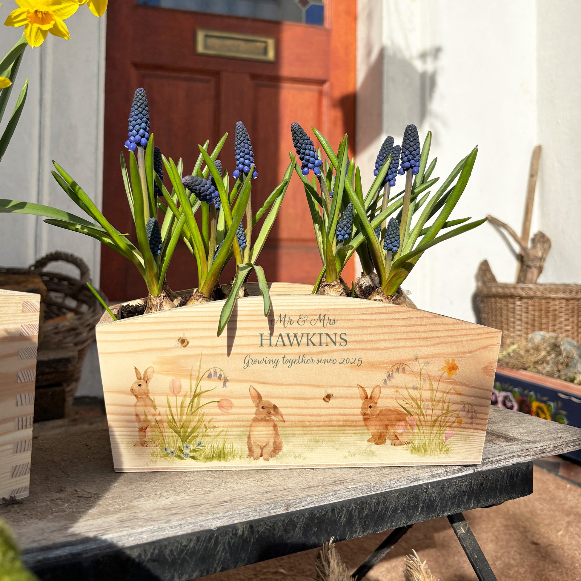 An arched wooden planter planter box showing a watercolour design of bunny rabbits among pastel spring flowers. The planter can be personalised with names and a selection of phrases, making it an ideal gift for any occasion.