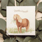 A greetings card laid flat on a table on top of dark green leaves. The greetings card reads Thanks a Bunch in brown text above a chestnut Shetland pony holding a bunch of flowers in a watercolour style.