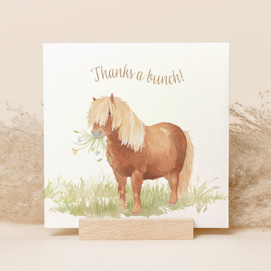 A greetings card reading Thanks a Bunch in brown text above a chestnut Shetland pony holding a bunch of flowers in a watercolour style.