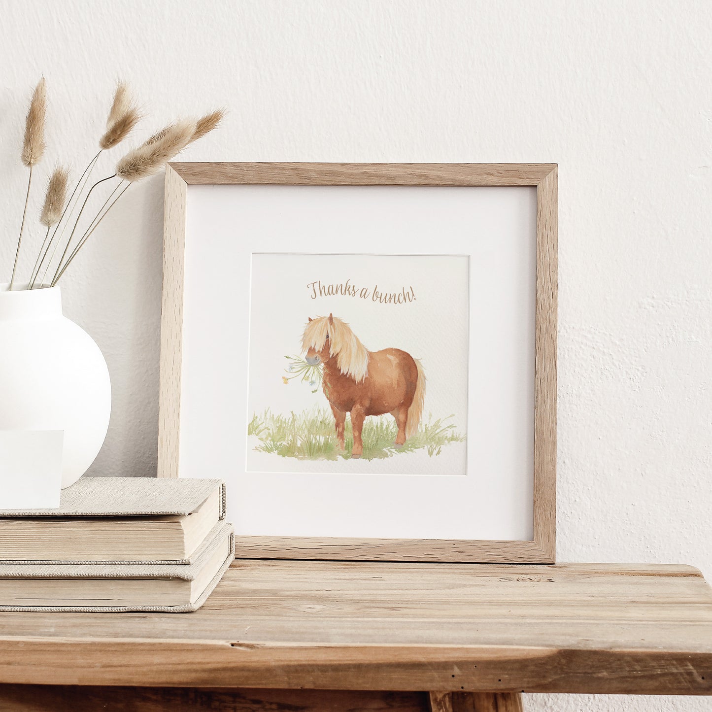 A greetings card displayed as an art print in a neutral coloured frame propped up on a bookshelf. The greetings card reads Thanks a Bunch in brown text above a chestnut Shetland pony holding a bunch of flowers in a watercolour style.