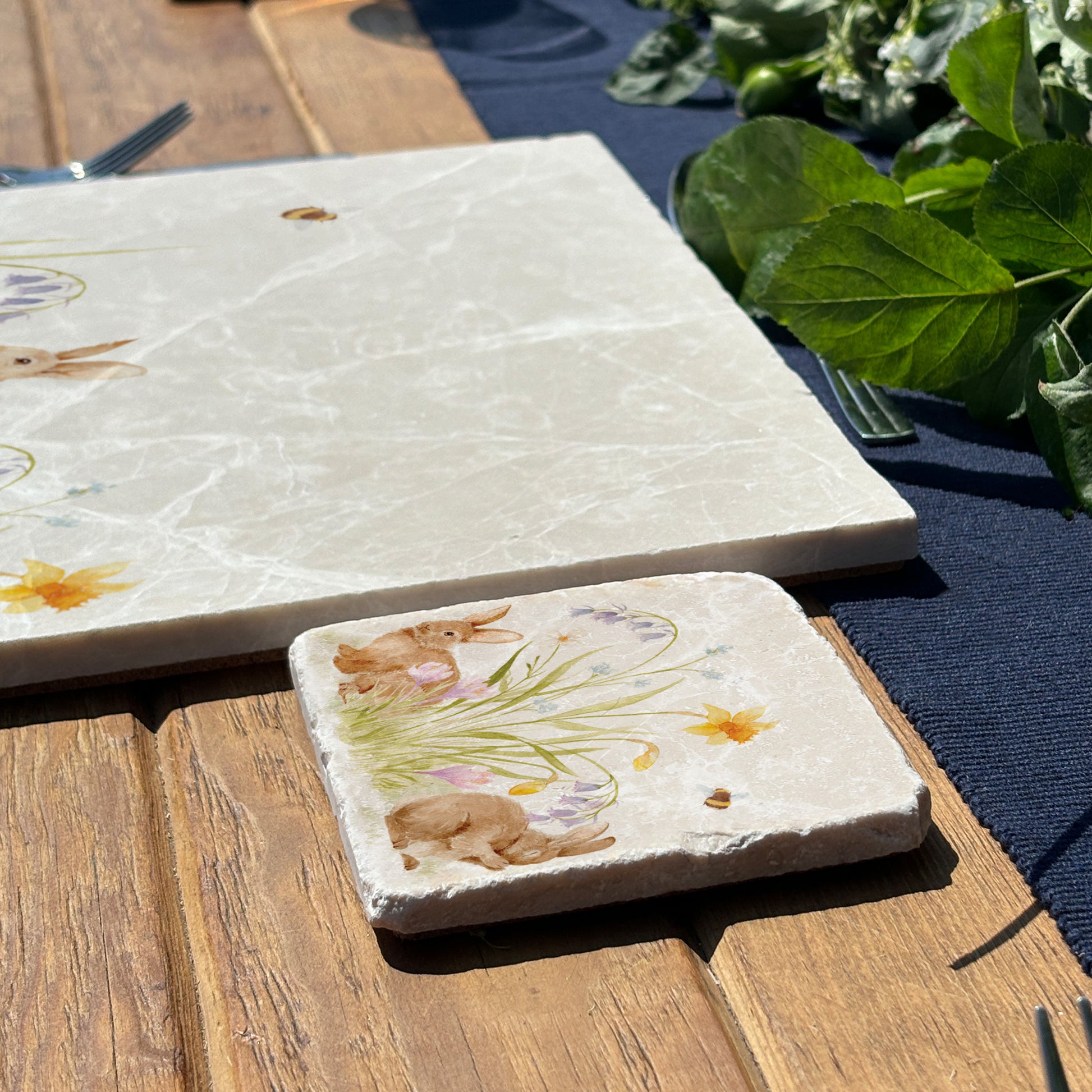A marble coaster set on a dining table. the coaster has a watercolour design of spring flowers and cute fluffy bunnies. 