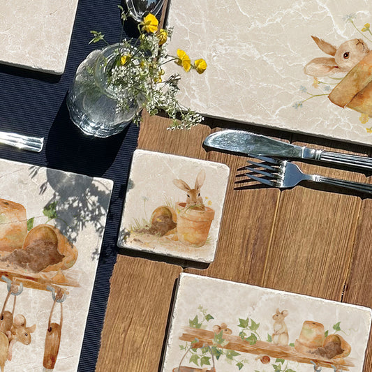 A marble coaster set on a dining table with placemats featuring a watercolour design of mischievous rabbits amongst the terracotta flowerpots and spring flowers.