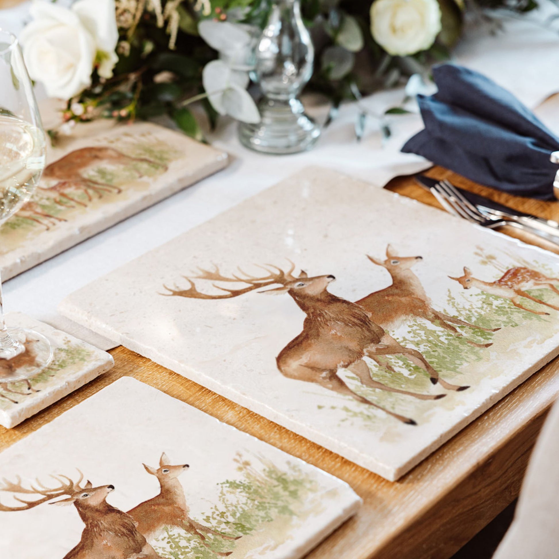 A wooden dining table set with cream marble placemats and coasters. The placemats are square and feature a family of deer in a watercolour style.