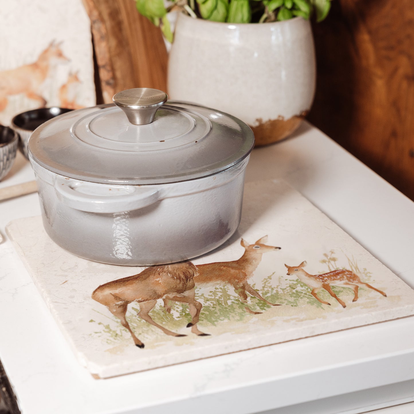 A large square marble platter featuring a watercolour design of a family of deer with a stag, doe and fawn. The platter is being used as a trivet.