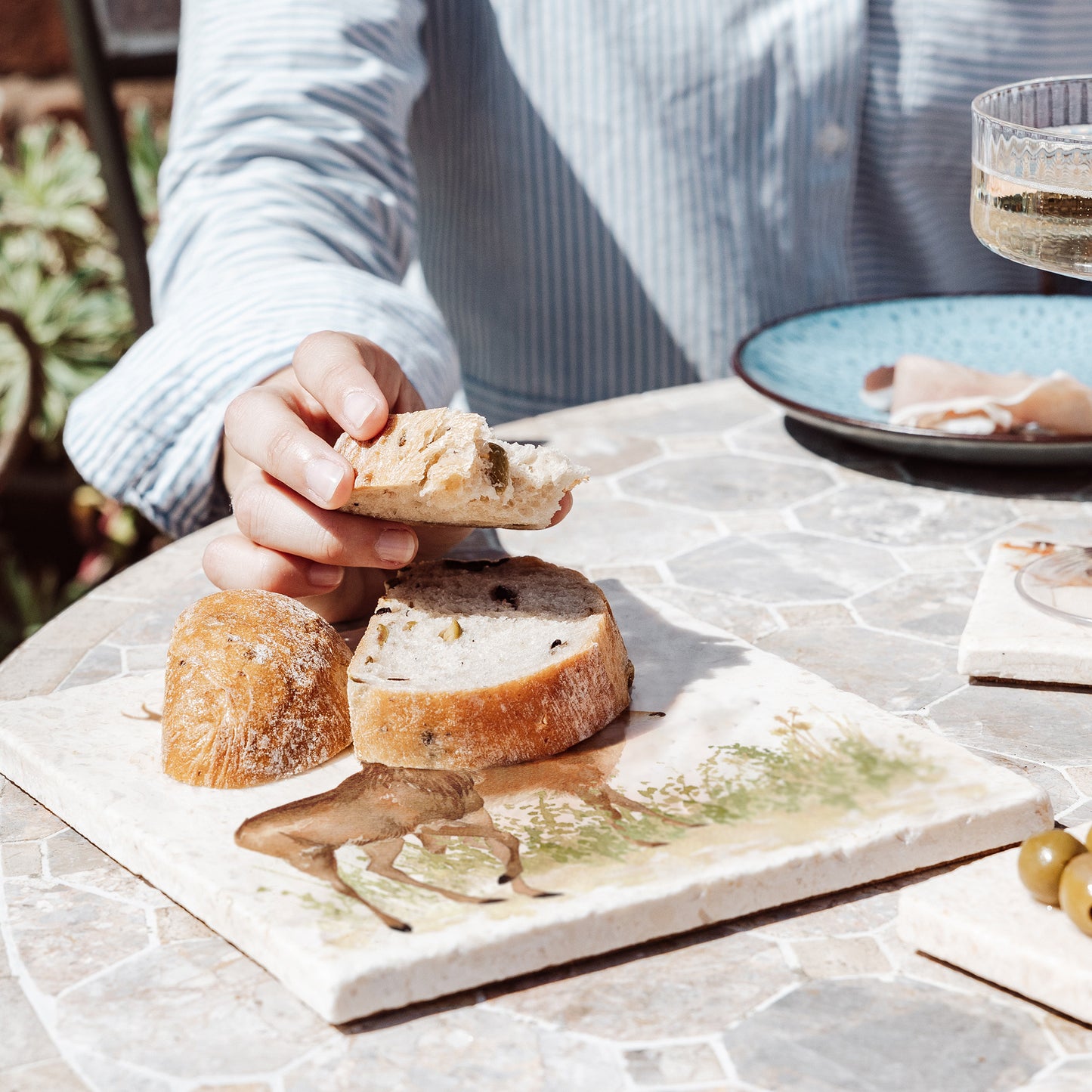 A woman's hand reaching to take a piece of bread from a medium marble bread board set on an al fresco dining table.