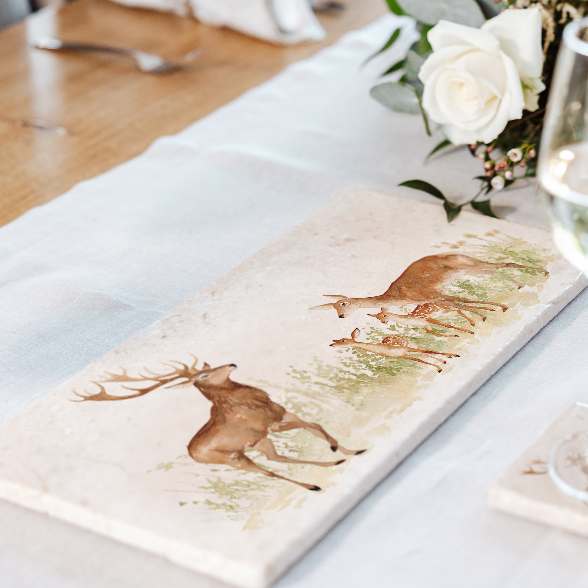 A luxurious dining table set with a rectangular marble sharing platter as a centrepiece. The platter features a watercolour design of a red deer family, including a stag, hind and two fawns.