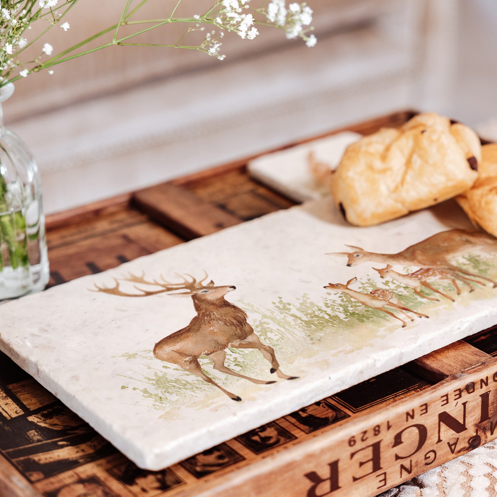 A rectangle marble board with a red deer family watercolour design featuring a stag, hind and two fawns. The platter is being used to serve pastries.