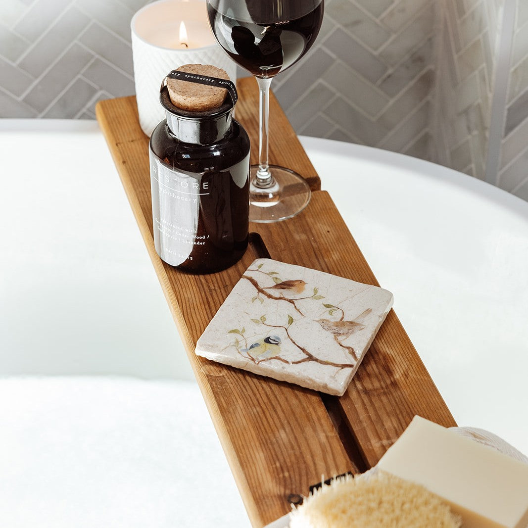 A square marble coaster on a wooden bath tidy above a bubble bath. The coaster features a British garden birds watercolour design.