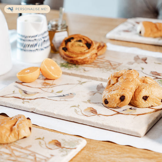 A breakfast table set with a marble sharing platter featuring British garden birds on branches in a watercolour style. The platter is being used to serve pastries and fruit.