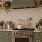 A styled kitchen scene with sage green cabinets, a gas stove, a cream pot, and a decorative tile mural depicting a rustic fence and sheep lined up on the backsplash.