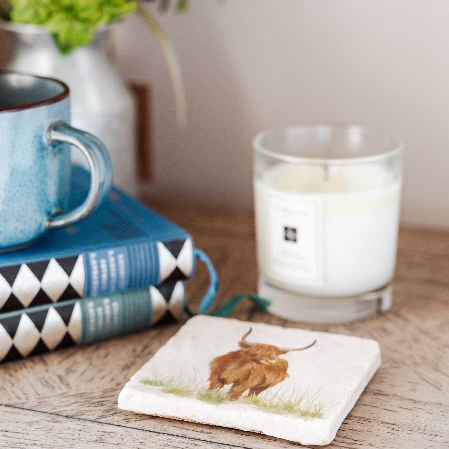 A square marble coaster set on a rustic wooden bedside table next to a candle, mug and books. The coaster features a watercolour design of a highland cow.