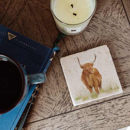 A square marble coaster featuring a watercolour highland cow design. The coaster is set on a rustic wooden bedside table.
