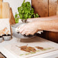 A large square marble platter with a highland cow design being used as a heat resistant trivet on a kitchen worktop. A woman is setting a hot casserole dish down on the platter.