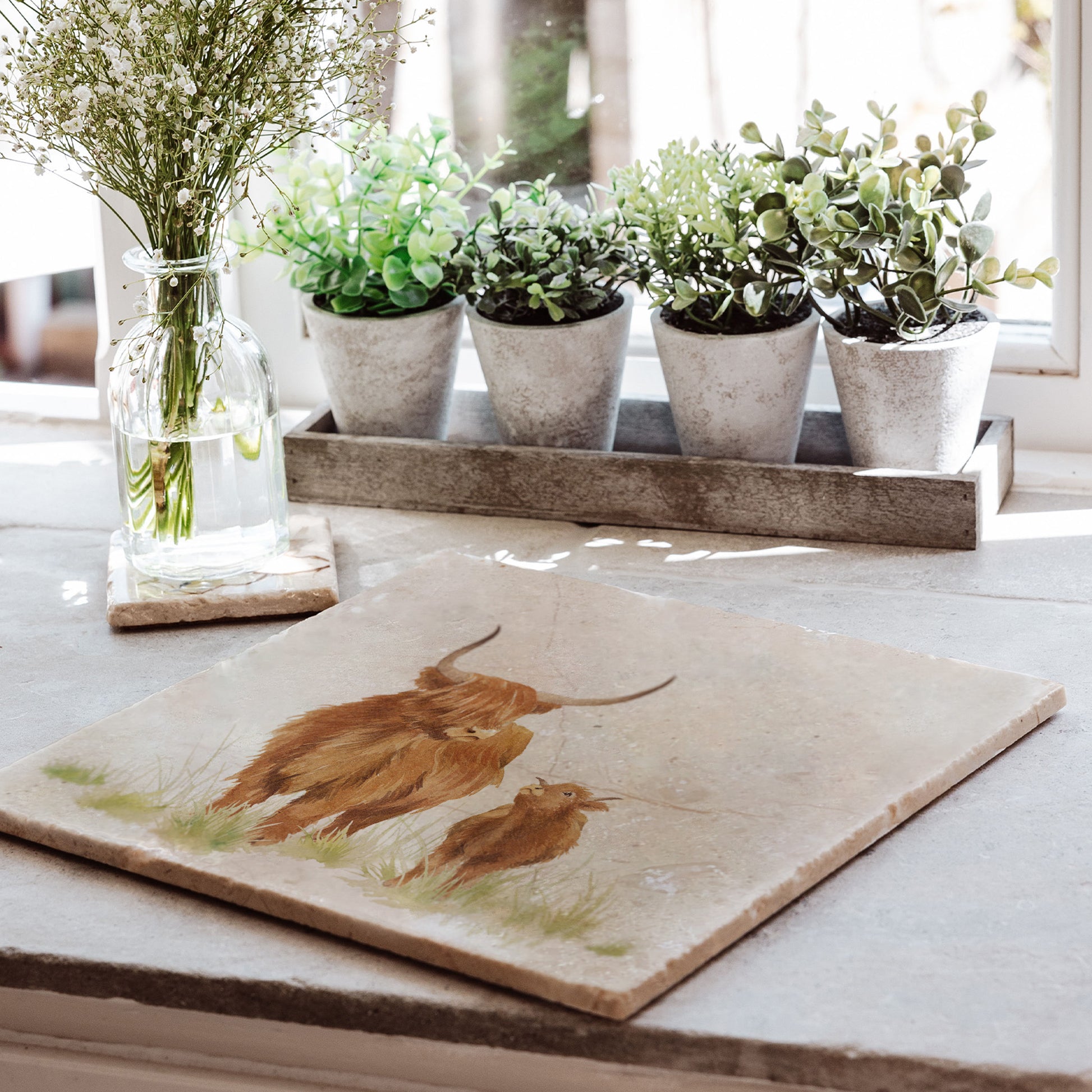 A large square marble platter placed on a sunny kitchen windowsill. The platter has a watercolour design of a highland cow and calf.