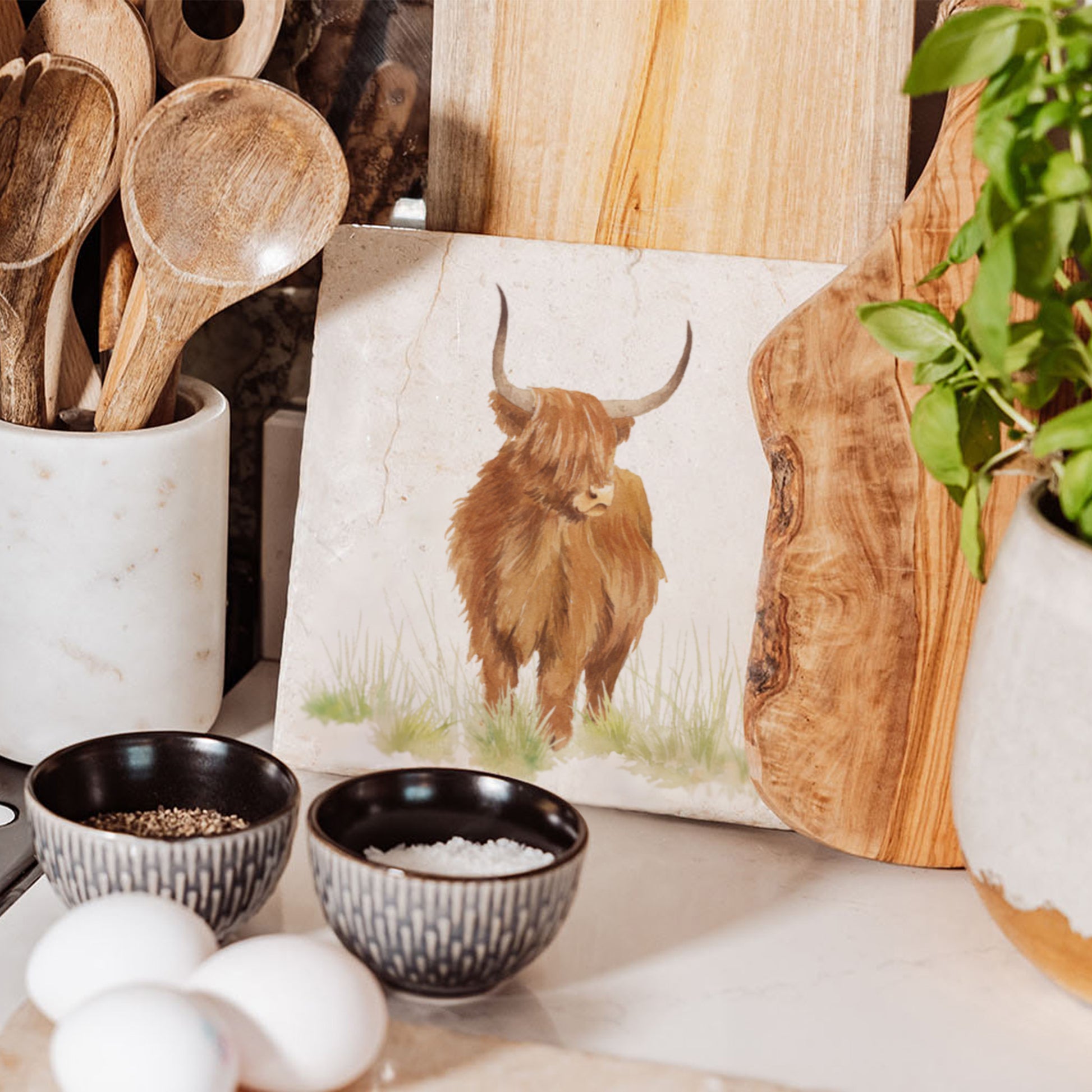 A medium square marble chopping board featuring a highland cow design, propped up on a kitchen worksurface. 