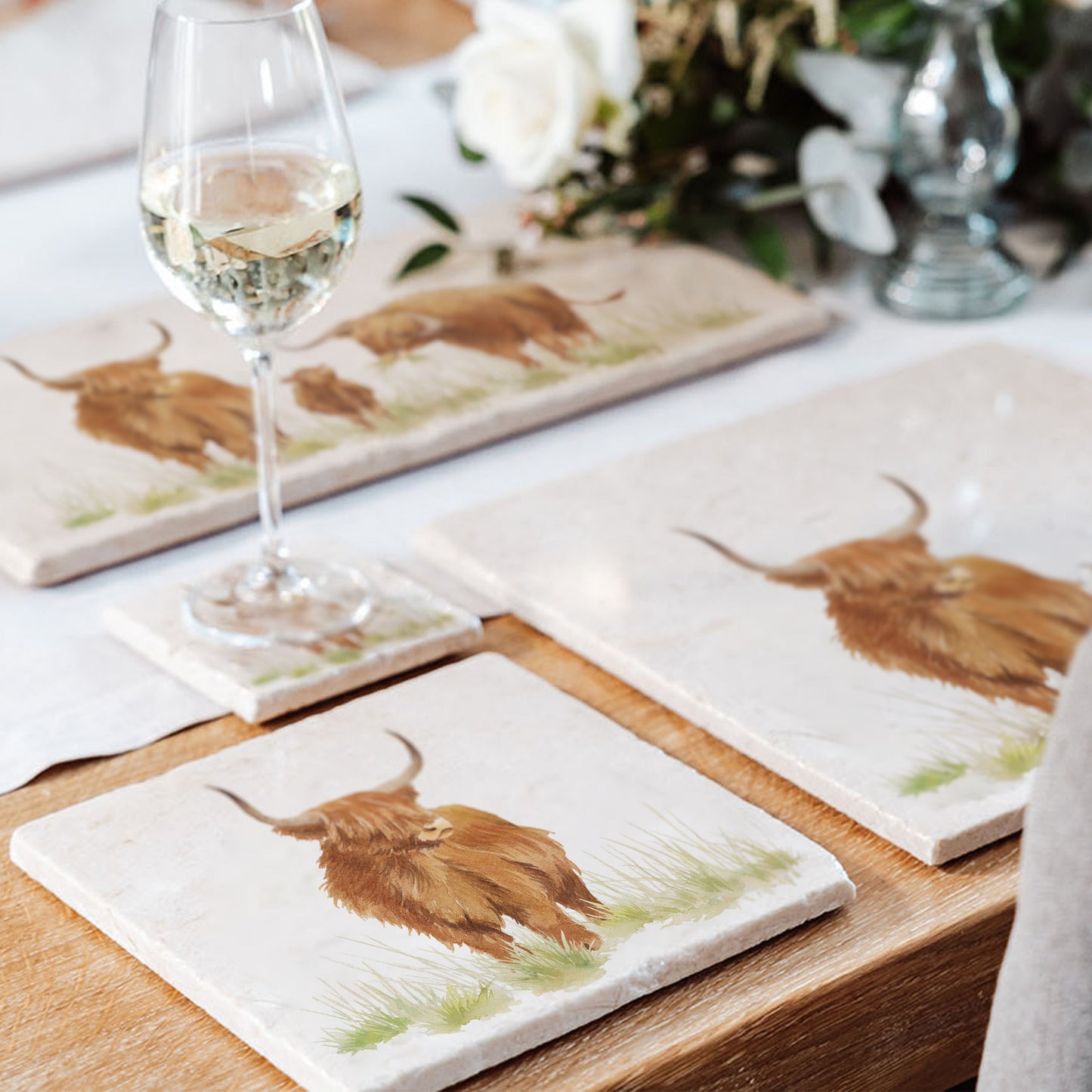 A wooden dining table set with cream marble placemats and coasters. The placemats are square and feature a daddy and baby highland cow