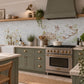 A decorative kitchen backsplash tile featuring a watercolour vintage-style illustration of birds perched on branches and a wooden fence, installed on a light grey tiled wall above a black stovetop in a styled kitchen setting.