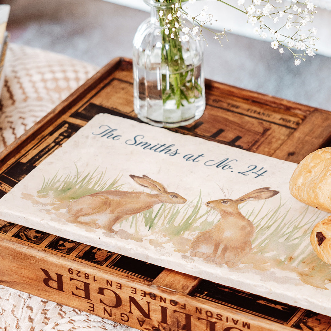 A personalised marble sharing platter featuring two kissing hares, the platter is placed on a coffee table to serve pastries. The personalised text reads 'The Smiths at No. 24'.